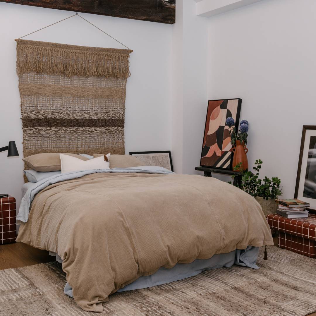 Bedroom with beige bedding, woven wall art, and abstract painting.