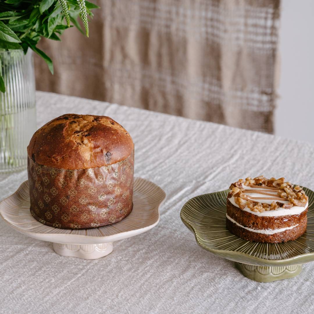 Two cakes on decorative stands on a textured surface with a plaid curtain in the background.