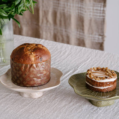 Two cakes on decorative stands on a textured surface with a plaid curtain in the background.