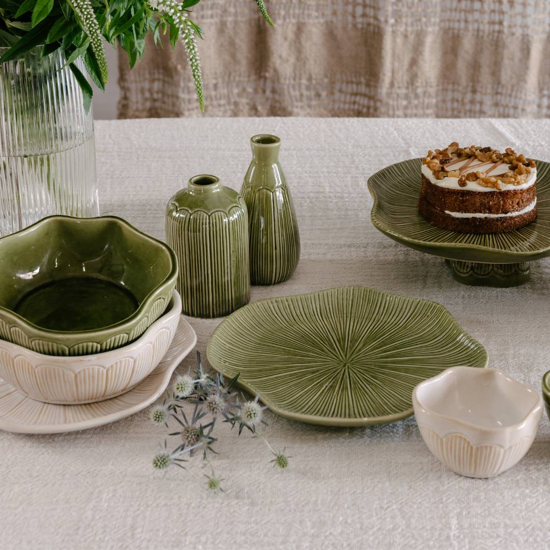 Green ceramic tableware on a light surface with a cake and plant in the background