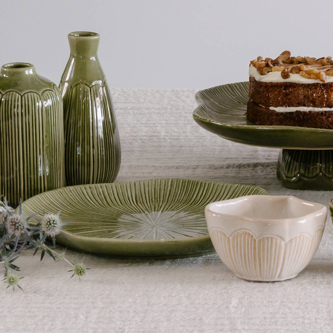 Green ceramic tableware including vases, plates, and a cake stand with a carrot cake on a neutral background.
