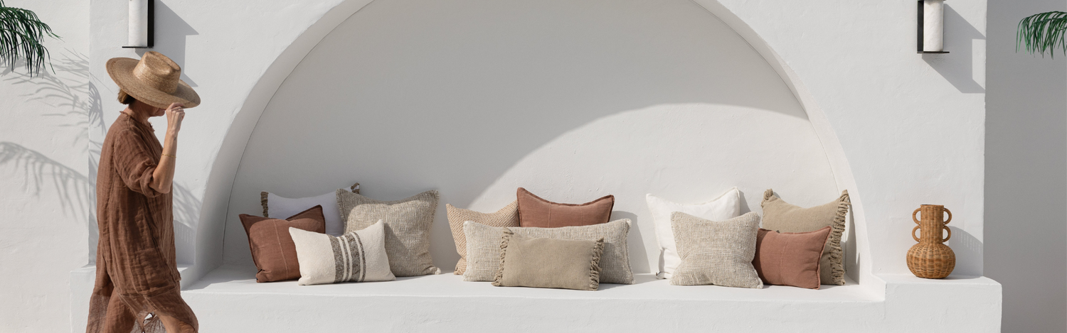 Woman in a long linen dress in clay colour and summer hat walking past to a white wall with a built-in bench featuring Eadie cushions from the Summer collection - Estella.