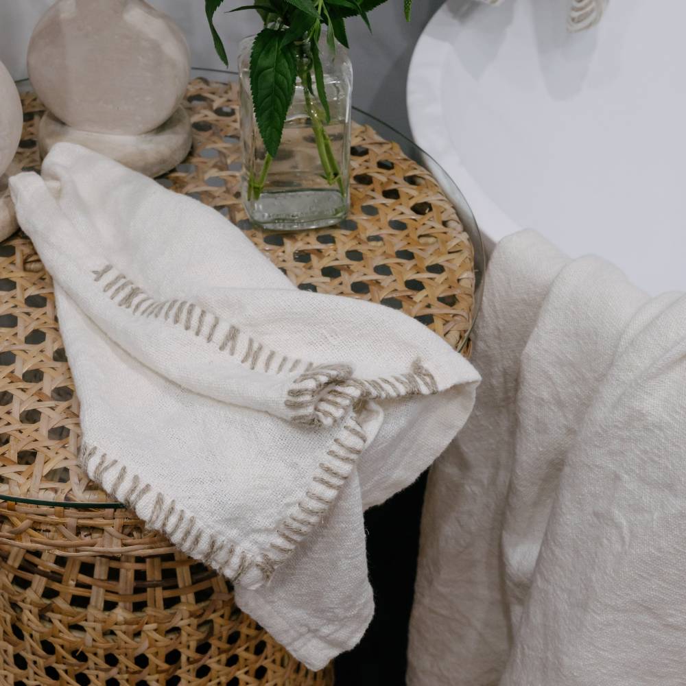 White towel with text draped over a woven stool with a vase of greenery in the background.